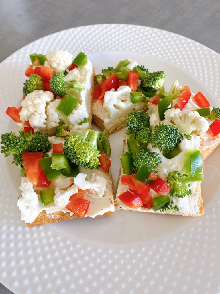 Sliced cold veggie pizza topped with broccoli, cauliflower, red and green bell peppers on a plate.